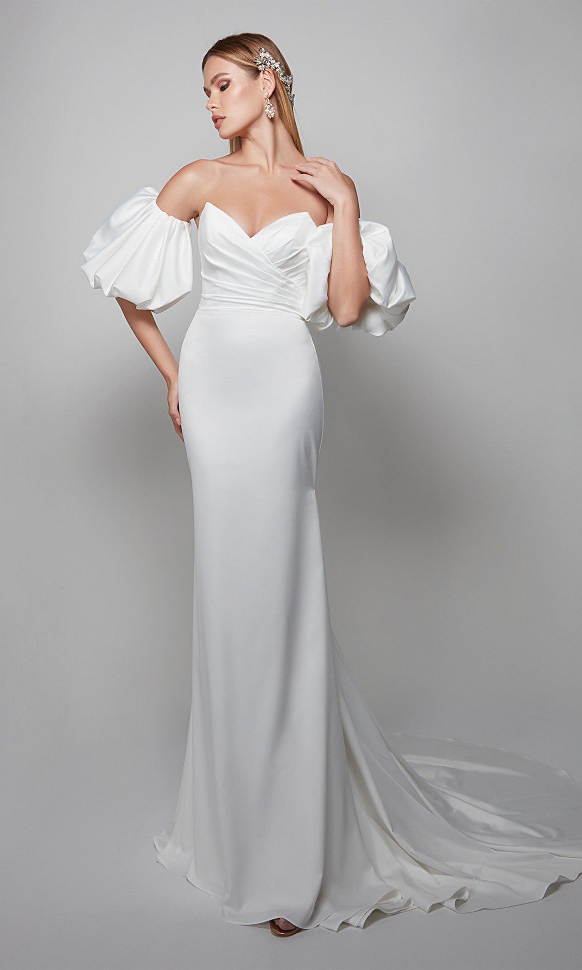 A woman wearing the Alyce 7056 long formal dress—white, off-the-shoulder with a sweetheart neckline, puffed sleeves, and fitted bodice—stands against a plain light background.